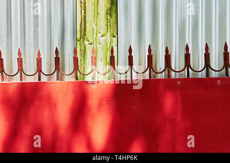 Détails de conception d'une grille métallique rouge avec des flèches soudé attaché au bord supérieur, que l'on voit contre un mur de feuilles de métal ondulé de couleur argent Banque D'Images