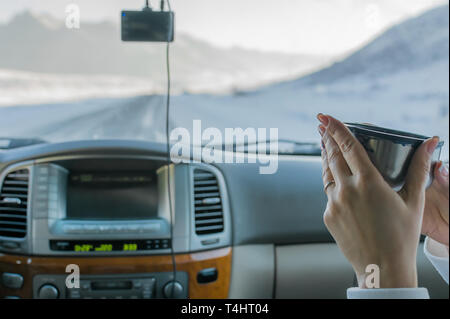 La main de la jeune fille dans le siège passager de la voiture maintenant du thé chaud à partir de la tasse de la thermos, vue sur la montagne à l'extérieur de la voiture de la route d'hiver Banque D'Images