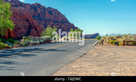 Tirer sur le pays route tournant autour de la montagne rocheuse rouge coloré sous ciel bleu. vert arbres et buissons, deux voies/ Banque D'Images