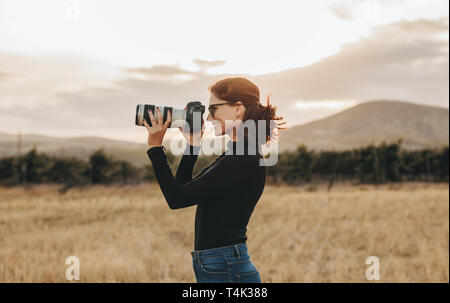 Vue latérale du jeune femme photographe prise de vue avec votre appareil photo reflex numérique à l'extérieur. Femme ayant une séance photo en extérieur. Banque D'Images