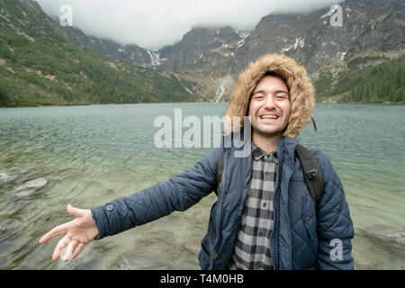 Heureux jeune homme par Lake dans les montagnes d'inviter à le rejoindre Banque D'Images