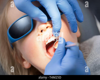 Dentiste dents de l'examen d'une petite fille dans une clinique dentaire pédiatrique à l'aide de miroirs inclinable. Enfant assis dans un fauteuil dentaire wearing sunglasses holding mo Banque D'Images