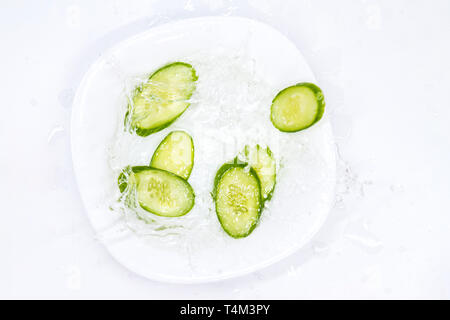 Les concombres et les projections d'eau dans une assiette sur un fond blanc, les ingrédients naturels Banque D'Images