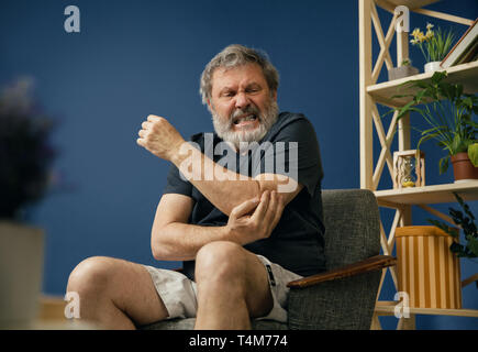 Insupportable. Vieux barbu assis sur la chaise et souffrant de douleur coudée sur fond bleu. Concept de la maladie, la perte de mémoire en raison de la démence, la santé, la dépression, le stress et la tristesse. Banque D'Images