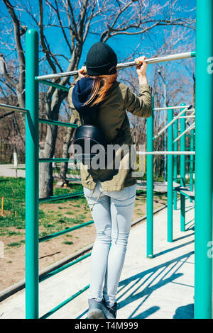 Young woman in casual vêtements avec sac à dos faisant tirer-se lève sur barre horizontale sur un terrain de sport aux beaux jours du printemps. Banque D'Images