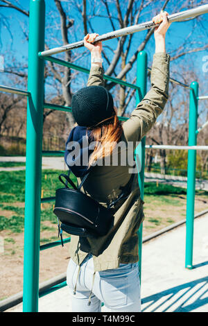 Vue arrière habillé en femme vêtements décontractés avec sac à dos faisant tirer-se lève sur barre horizontale sur un terrain de sport aux beaux jours du printemps. Banque D'Images