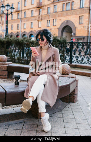 Vue de côté de belle jeune femme à lunettes en utilisant son smartphone et à l'écart avec le sourire tout en restant assis à l'extérieur Banque D'Images