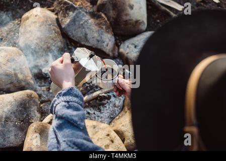 Traveler woman in hat verse lui-même boisson chaude dans les montagnes près de feu . Jeune femme assis et tenant une tasse de café après la randonnée. Locations Banque D'Images