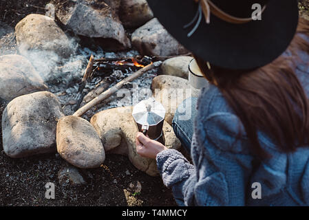 Traveler girl in hat est titulaire d'un café chaud dans une forêt près de feu. Femme assise et tenant une tasse de café après une randonnée.concept Trekking Banque D'Images