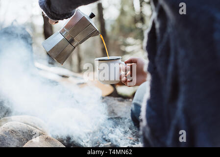 Les mains des femmes pour lui-même du café chaud dans une forêt près de feu. Fille assise et tenant une tasse de café après la randonnée. Concept Aventure Banque D'Images