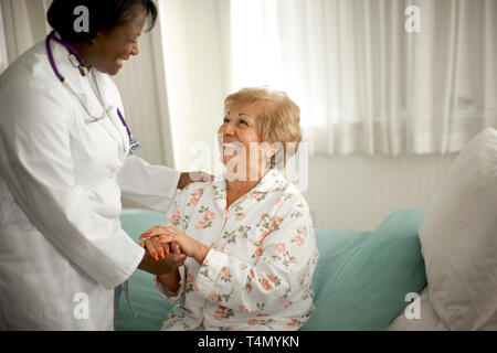 Smiling senior woman holding la main d'une femme médecin alors qu'elle se trouve sur le bord d'un lit à l'intérieur d'une chambre. Banque D'Images