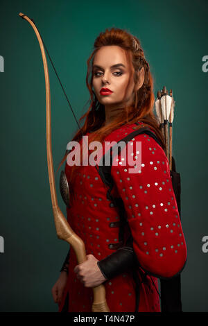 Belle femme guerrière tenant en main, l'arc avec des flèches derrière le dos, portant en rouge tunique moyenâgeuse. Courageux, femme sérieuse avec du gingembre des cheveux, des lèvres et des oreilles elf looking at camera, posant. Banque D'Images