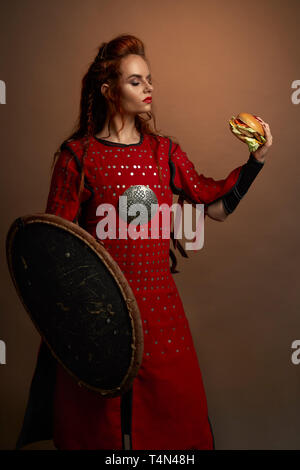 Belle femme warrior holding délicieux hamburger à la main, le regardant. Sérieux, modèle courageux de porter à l'époque médiévale robe rouge, tenant debout, bouclier et posant. Banque D'Images