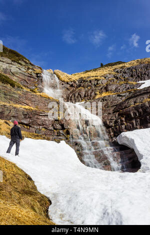 Le Randonneur avec Chapeau hiver pourpre debout devant le grand scenic, belle cascade tombant d'une falaise rocheuse de rouge sous la neige Banque D'Images