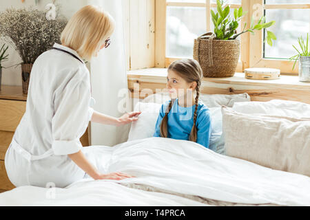 Mettre un kit pour un lit. Charmant agréable à poil court costumes de pédiatre dans un manteau blanc mettre petit mignon brun fille malade au lit. Banque D'Images