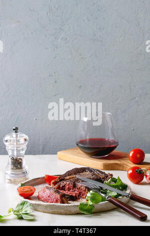 Tranches de rôti grillé à point Pavé de boeuf servi dans une plaque en céramique avec champ vert salade, tomates cerise, couverts, poivre, verre de vin rouge sur Banque D'Images