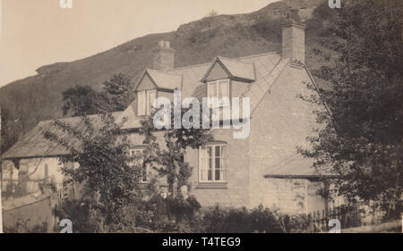 Vintage Carte postale photographique montrant une maison historique de l'Intérieur britannique dans un emplacement rural au pied d'une colline. Couple se tenait dans le jardin de la propriété. Banque D'Images