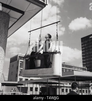 Années 1960, historiques, deux vitres debout dans un palan en béton avec godet, suspendue au-dessus, à la hauteur à l'immeuble de bureaux qu'ils auront à nettoyer à la main, Londres, Angleterre, Royaume-Uni. Banque D'Images