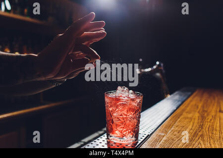 Les mains de barman en saupoudrant l de jus dans le verre à cocktail rempli de boisson alcoolisée sur le fond sombre. Banque D'Images