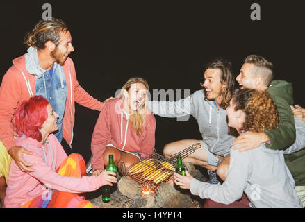 Happy friends camping ensemble et la cuisson du maïs à l'extérieur nuit - Les jeunes s'amuser et rire autour de feu Banque D'Images