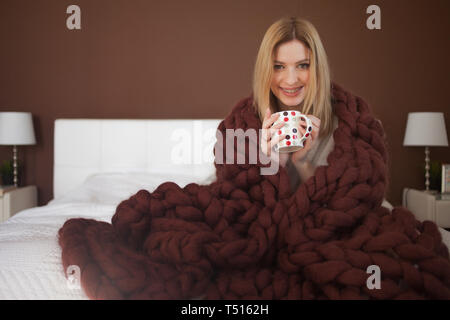 Happy young woman in Bathrobe sitting on bed enveloppés dans de grandes et de moelleux plaid marron. Belle fille dans une couverture chaude avec une tasse de boisson. Et chaud coz. Banque D'Images