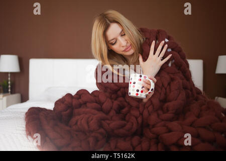 Happy young woman in Bathrobe sitting on bed enveloppés dans de grandes et de moelleux plaid marron. Belle fille dans une couverture chaude avec une tasse de boisson. Et chaud coz. Banque D'Images