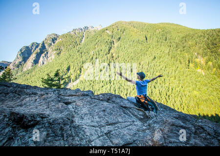 Randonneur une femme assise sur le bord d'une falaise rocheuse à la recherche jusqu'à une montagne avec les bras dans la célébration, si peu de Trail, Washington, USA. Banque D'Images