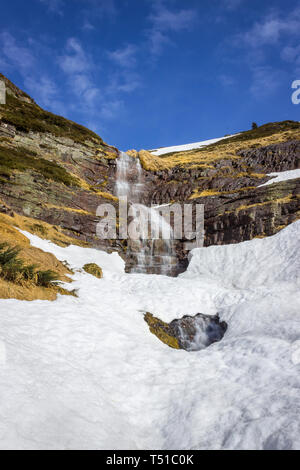 Grand scenic, belle chute d'eau tombant d'une falaise rocheuse de rouge sous la neige pendant la période de transition entre l'hiver et au printemps Banque D'Images