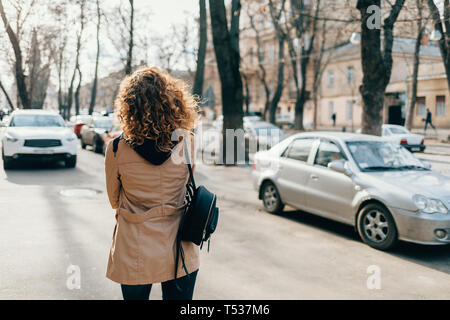 Retour voir jeune femme avec des cheveux bouclés portant trench-coat beige et son sac à dos en descendant dans la rue près de la ville avec des voitures de route. Banque D'Images