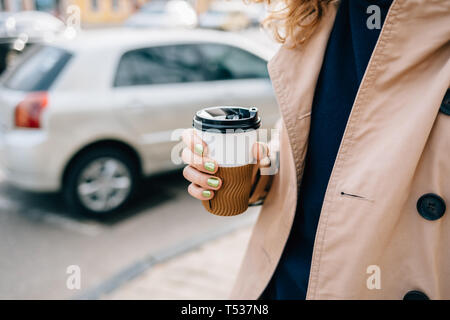 Café chaud pour aller dans la main de femme, close-up. Jeune femme tenant un gobelet jetable de boisson chaude walking on city street près de road avec des voitures à sp Banque D'Images