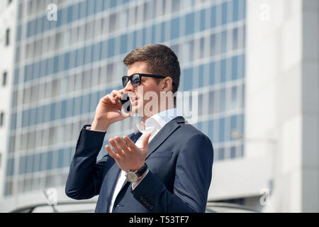 Young businessman wearing sunglasses debout près de conversation sur smartphone voiture ayant des gestes expressifs expliquant problème close-up Banque D'Images