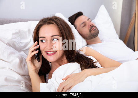 Smiling Young Woman talking On Smartphone en dehors de son mari sur le lit dans la chambre à coucher Banque D'Images