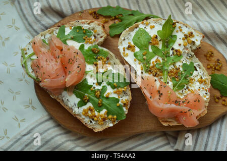 Des sandwiches au saumon avec du fromage à la crème, les graines de moutarde, l'huile d'olive, roquette sur pain de seigle grillé, servi sur la planche de bois. La mode en bonne santé l'alimentation. Banque D'Images