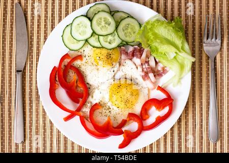 Poulet frit délicieux et frais oeufs frais avec du poivre rouge, du bacon, de la laitue et le concombre. Le plat est sur une plaque blanche avec un couteau et fourchette. Banque D'Images