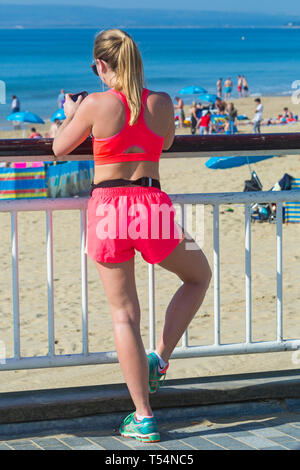 Bournemouth, Dorset, UK. 21 avr 2019. Météo France : la canicule se poursuit avec le temps ensoleillé et chaud, en tant que chef de la plage au bord de la mer pour profiter de la chaleur et du soleil à plages de Bournemouth pour les vacances de Pâques - le milieu de la matinée et les plages sont déjà obtenir paniers, comme sunseekers arriver tôt pour obtenir leur espace. Young woman standing on pier looking at mobile phone. Credit : Carolyn Jenkins/Alamy Live News Banque D'Images