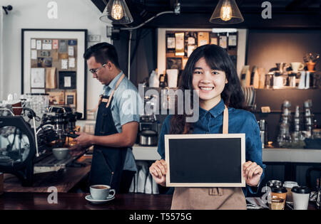 Asian female barista porter jean apron holding blank blackboard menu café au comptoir bar avec collègue avec Smile, cafe service concept,business Banque D'Images