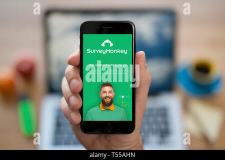 Un homme se penche sur son iPhone qui affiche le logo Survey Monkey (usage éditorial uniquement). Banque D'Images
