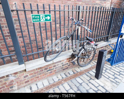 Une dame noire ou d'une femme's bike verrouillé sur certaines rampes d'acier en face de mur de briques, sur une route pavée Banque D'Images