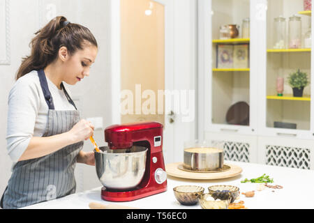 Jeune femme pâtissier bat la crème dans un bol en métal rouge dans un mélangeur électrique. Le concept de pâtisserie maison, la cuisine. Banque D'Images