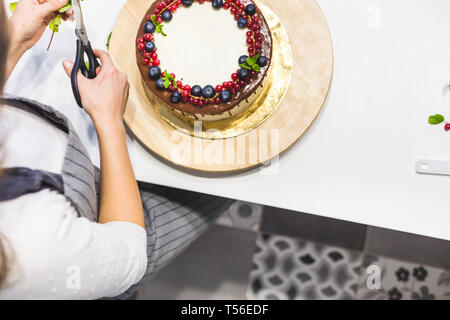Pâtissier décore avec des baies un biscuit gâteau au chocolat blanc et crème. Cake se dresse sur un support en bois sur un tableau blanc. Le concept de Banque D'Images