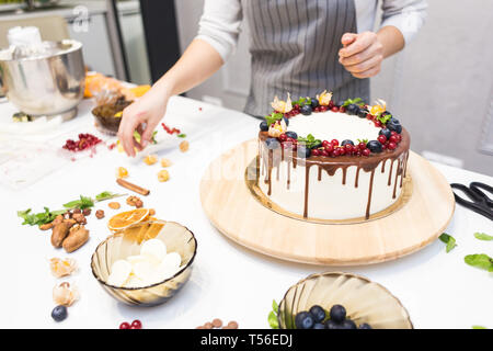 Pâtissier décore avec des baies un biscuit gâteau au chocolat blanc et crème. Cake se dresse sur un support en bois sur un tableau blanc. Le concept de Banque D'Images