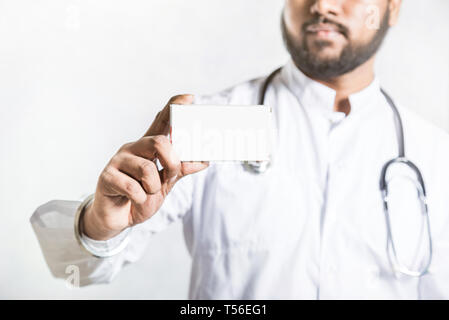 Beau jeune médecin dans une robe blanche avec un stéthoscope détient sur une main tendue un paquet blanc vierge fort pour l'emballage de comprimés. Des maquettes. Banque D'Images