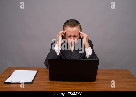 Mature Asian businessman sitting avec table en bois contre gray Banque D'Images