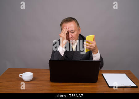 Mature Asian businessman sitting avec table en bois contre gray Banque D'Images