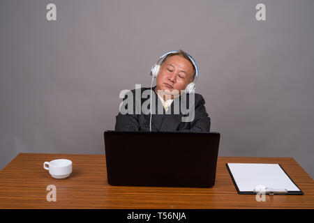 Mature Asian businessman sitting avec table en bois contre gray Banque D'Images