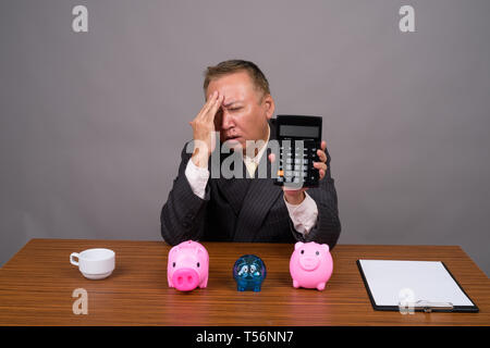 Mature Asian businessman sitting avec table en bois contre gray Banque D'Images