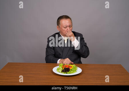 Mature Asian businessman sitting avec table en bois contre gray Banque D'Images