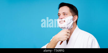 Photo de heureux homme brunet avec rasoir droit en robe blanche sur fond bleu blanc en studio. Un espace réservé au texte Banque D'Images