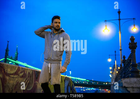 Un beau jeune sportsman debout dans les lumières de la ville au bord de la rivière dans la nuit avec un pont derrière lui Banque D'Images