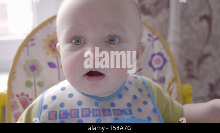 Beau petit bébé garçon assis à la table des enfants dans le bleu bib, regarder dans la caméra et babillant voir mo lente en vidéo 4K Banque D'Images